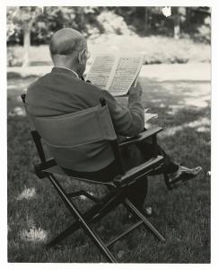 Older man seated in folding chair on lawn facing away, legs crossed, wearing glasses.
