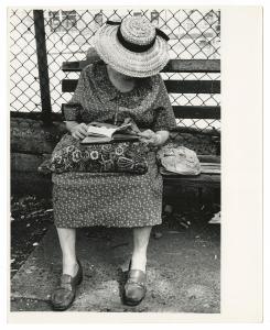 Older woman sitting on park bench in front of chain link fence, wearing dress and hat, pillow in lap.