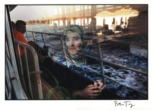 Photo of a woman in a blue head scarf on a ferry, with the sea and sky reflected off the window she is looking through