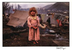 A young refugee girl barefoot in a pink dress at a Kurdish refugee camp, with a makeshift tent made from a rug in the background