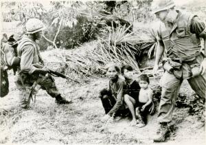 Black and white photo of 2 children and a woman seated on the ground with a US soldier on either side of them. The one on the left points a rifle at the civilians.