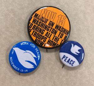 Image of 3 VIetnam protest buttons. Two blue buttons have a white dove. The largest button is orange with black bold letters.