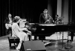 B X W photo of jazz band with woman in formal dress playing piano, man playing guitar, a man playing saxophone with male drummer in background