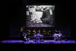 Color photo of 10 men playing music with a photograph of a man at a grave above them.