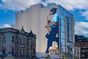 Color photo of mural showing Buck Hill, a man playing saxophone and wearing letter carrier uniform with his bag at bottom of mural; DC row houses in foreground and office buildings in background.