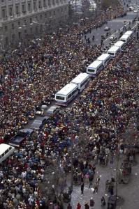 Color photo of parade with white buses and black cars in center and crowds looking on 