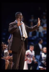 Color photo of tall black man with palms facing forward and in brown suit wearing a white towel over his left shoulder