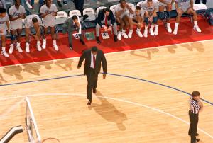Overhead color photo of Coach John Thompson walking on basketball court with his players applauding; red boundary around basketball court