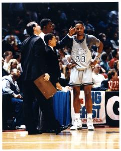 Color photo of Basketball center on sideline with coach tapping his head; other coaches in foreground; Big East Conference banner in background