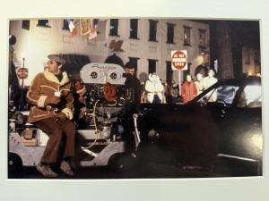 Film director William Friedkin sitting next to a film camera, with cast and bystanders in the background during the night
