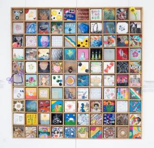 One hundred five-inch by five-inch squares hung in a rectangular wood frame. Each square is distinctly different in style, with a variety of colors and textures. The media on display include painting, collage, photography, laser cut wood, photography, text, and hand sewing.