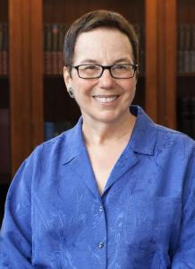 Harriette Hemmasi wearing a blue blouse and glasses standing in front of bookcases
