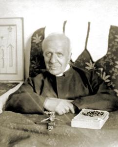 Benedict Sestini, and older priest with balding white hair, sits in an upholstered chair with a crucifix, book, and rosary on a table in front of him