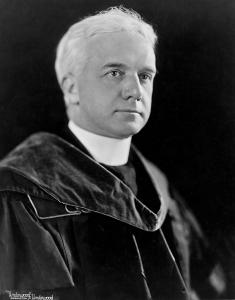 Coleman Nevils, a middle aged priest with white hair wearing academic robes and a tall white collar
