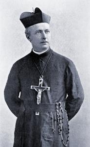 Francis Barnum, a middle-aged priest wearing dark robes, a Jesuit biretta, and a large crucifix around his neck, stands wih his hands behind his back
