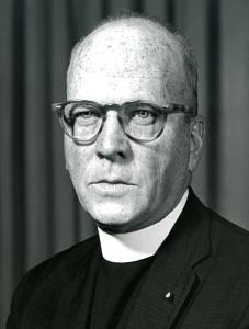 Geeard Yates, and older priest with freckles and a receding hair line wearing thick rimmed glasses, a dark coat and a tall white collar