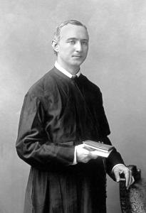 Henry Shandelle, a young priest with short light hair, stands with a book in his right hand and his left hand resting on the back of a chair