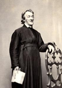 James Curley, standing, wearing black robes, holding a book in his right hand and resting his left hand on the back of a chair