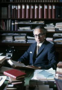 Joe Jeffs holds an open book behind a desk covered with stacks of books and paper, wearing a blue suit and tie and thick rimmed glasses