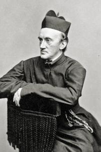 John Sumner, a priest in dark robes and a Jesuit biretta, sits with his crossed arms resting on the back of a chair