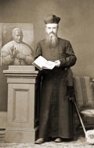 Joseph Finotti, a priest with a long beard and moustache wearing long black robes and a Jesuit biretta holds an open book and stands next to a painting of Pope Pius IX on a pedestal