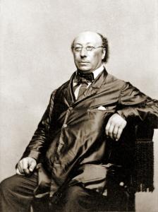 Leonard Nota, a balding man with dark curly hair and glasses with octagonal frames, sits in a chair for a portrait wearing a long dark jacket and western style bow tie