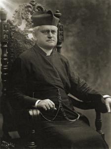 Michael O'Brien, a middle aged priest with short gray hair in dark robes and a Jesuit biretta sits on a wooden chair with an intricately carved back