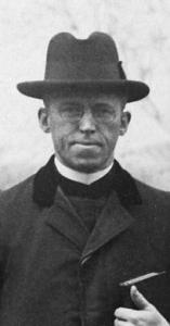 Philip Burkett, a middle aged priest in glasses holds a book under his left arm and wears a coat and Homburg style hat