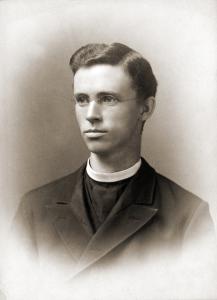 A soft vignette portrait photograph of Timothy Brosnahan, a young priest with dark wavy hair and glasses wearing a dark jacket and white collar 