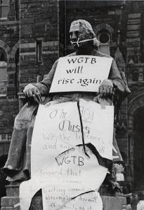 Black and white photograph showing the John Carroll statue gagged and holding a sign reading "WGTB will rise again"