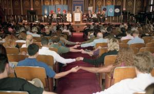 Color photograph taken from the back of Gaston Hall looking toward the stage. It shows seated attendees holding hands across the aisle, with  participants on the stage seen in the background.