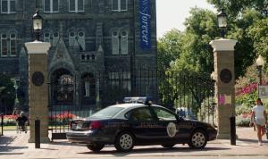 Color photograph showing a GU Department Public Safety police car parked in front of closed gates at the main University entrance. The north end of Healy Hall with a blue "Georgetown Forever" banner on it, can be seen in the background