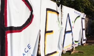 Color photograph showing a row of decorated bedsheets spelling on the word "Peace" hung on a stone wall.