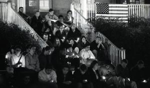Black and white photograph showing students who are holding candles sitting in the Quadrangle on the steps of Old North.