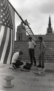 Black and white photograph showing three people on a roof top examining the damaged antenna which has a U.S. flag attacked to its tip.
