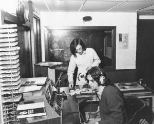 Black and white photograph of radio studio with a program host wearing headphones, sitting at a microphone, and a person standing behind him, assisting