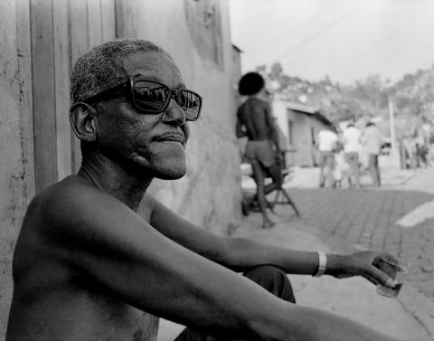 Black and white photograph of a dark skinned man in profile, shoulder length. He wears large sunglasses and rests against a well with one arm on his knee.