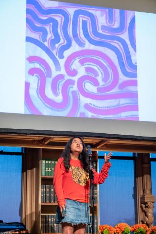A performer with long black curly hair wearing a red jumper with a tiger on it, and a denim skirt. There is a graphic of pink and blue swirly, thick lines projected at the back
