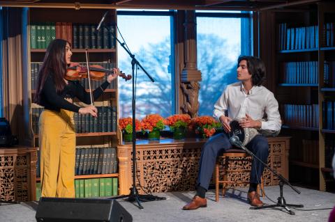 Two performers on stage one with long brown hair wearing yellow pants and a black shirt playing a violin. The other with short black hair wearing blue slacks and a white dress shirt playing a darbuka