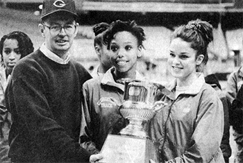 Senior associate athletic director Joe Lang presents the Big East women's track and field championship trophy to student athletes Kari Bertrand and Steffanie Smith. The three are standing together in a black and white image and smiling, looking away from the camera.