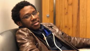 A Black male student with glasses sits back in a chair.