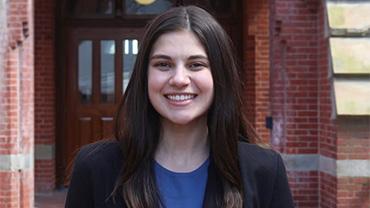 A white female student with brown hair in a suit in front of a brick building.