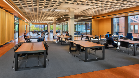 View of the renovated Pierce Reading Room, featuring long, polished wooden study tables lined with classic reading lamps, high ceilings, large windows, and students studying, reading, and talking. View of two large group study rooms in the background.