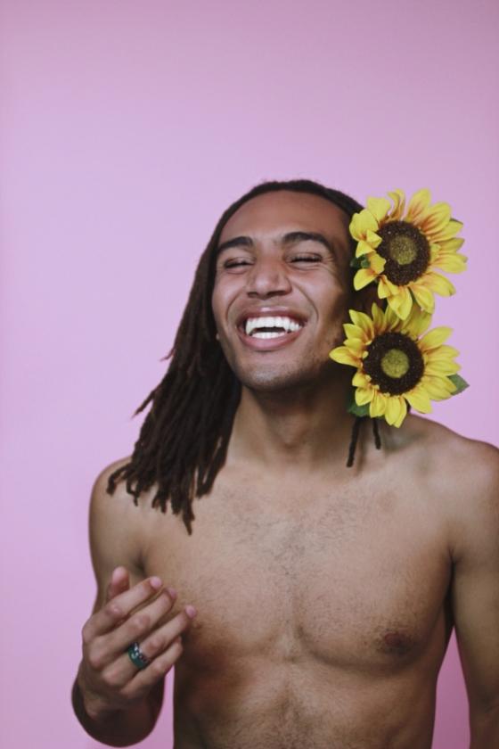 A shirtless black man with dreadlocks and two sunflowers tucked over his left ear smiles with his eyes closed 