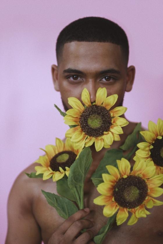 A shirtless black man stares at the camera while the bottom of is face is hidden behind four yellow sunflowers that he holds in his right hand