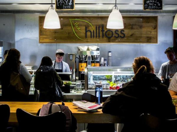 Image of the counter at The Hilltoss. A Corp member is facing the camera and assisting a customer who's back is to the camera. The Hilltoss sign is visible on the wall behind the Corp member.