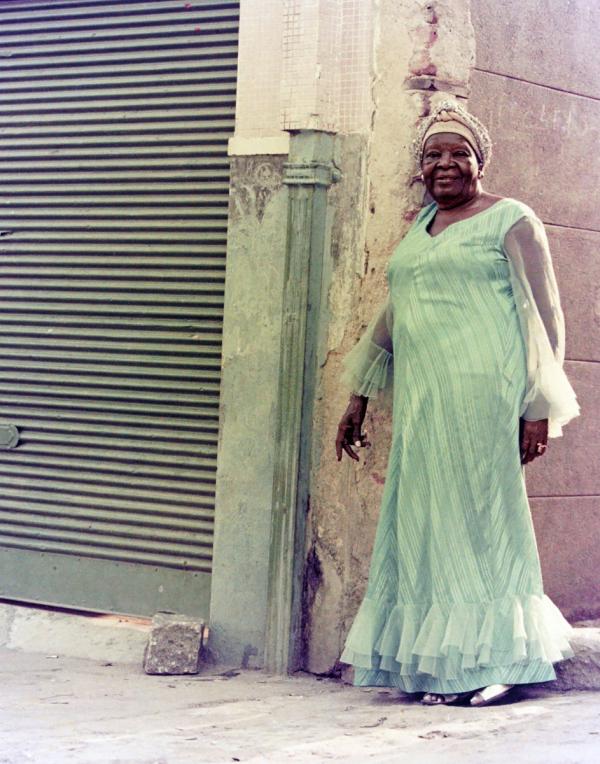Color photographic portrait of woman standing in front of building corner. She wears a green long sleeve dress which echoes the green tint of the closed shop or garage around the corner..