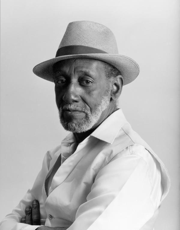 Black and white portrait of an old man wearing a hate and a crisp white suit. He is standing semi profile and from the chest up so that only a little bit of his crossed arms are visible.
