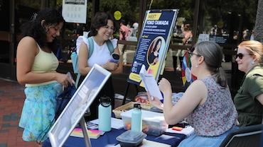 Two women standing in front of a table and two women sitting at the table. There are posters and flyers around the table.