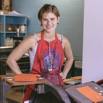 woman wearing a messy orange apron standing by a printing press